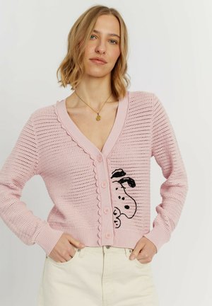Donna che indossa un cardigan lavorato a maglia rosa chiaro con bordi smerlati e disegno di cane cartoon nero, abbinato a pantaloni chiari, mani nelle tasche.