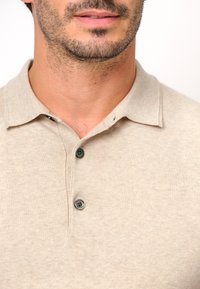 Beige polotröja i mjukt tyg, med klassisk krage, två mörka knappar och en liten tråddetalj nära kragen.