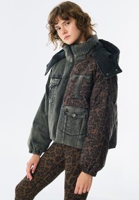 Jeune personne portant une veste rembourrée à imprimé léopard gris et marron avec un grand patch en forme de "T", associée à des leggings assortis à imprimé léopard.