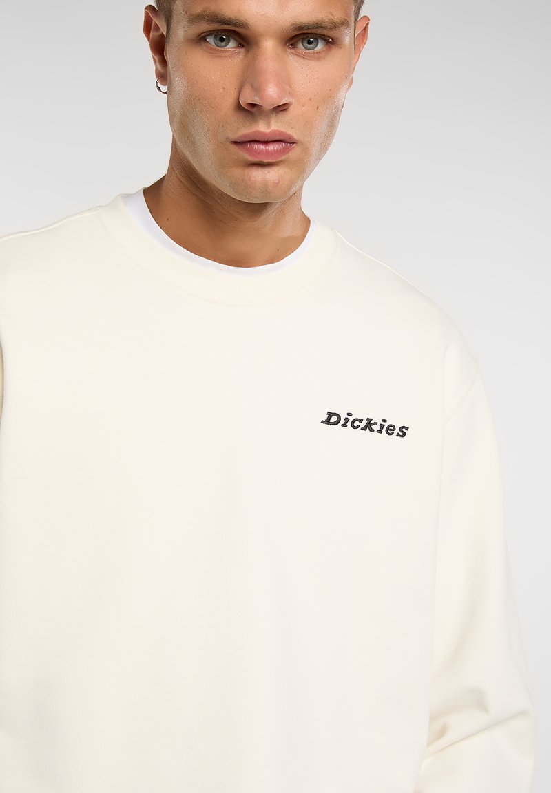 Sudadera color crema con un ajuste relajado, cuello acanalado y logo "Dickies" bordado en negro en el lado izquierdo. Textura suave.