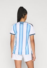 Mulher com cabelo trançado, vestindo uma camisola desportiva listrada verticalmente em azul e branco com "1893" nas costas e calções brancos.