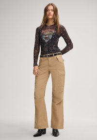 Zwarte longsleeve top met tijgerafbeelding en kant patroon, gecombineerd met tan cargobroek met zakken en een zwarte riem. Zwarte enkellaarsjes maken de look af.