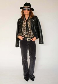 Veste de moto en cuir noir portée sur un chemisier à motifs, assortie à un pantalon en velours côtelé foncé évasé et des escarpins noirs à bout pointu. Accessoirisée avec un chapeau et des lunettes de soleil.