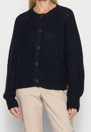 Femme portant un cardigan en maille épaisse bleu marine avec des boutons et un pantalon beige, montrée du cou jusqu'à mi-cuisse sur un fond uni.