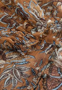 Hnedá látka s kvetinovým vzorom, ktorá obsahuje svetlomodré botanické ilustrácie, nafrkavané detaily a zlaté metalické prúžky. Zahrnuje akcenty v podobe gombíkov.