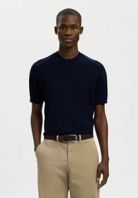 Pull en maille bleu marine à manches courtes avec un col rond, une texture lisse et une coupe ajustée, associé à un pantalon kaki clair et une ceinture tressée marron.