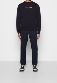 Marinblå sweatshirt med logobroderi i rött, grönt och orange. Kombinerad med mörka byxor och marinblå sneakers med cremefärgade detaljer och bruna sulor.