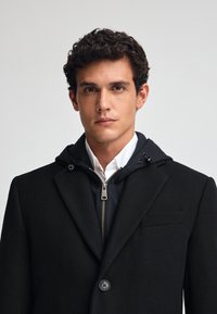 Svart ullblandad kappa med en dragkedjeförsedd huva, som har en enkel knappstängning, skarpa slag och en bröstficka.