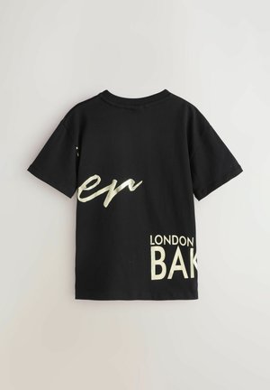 Sort bomulds T-shirt med metallisk gul grafisk tekst "en" og "LONDON BAK" på ryggen. Korte ærmer og design med rund hals.