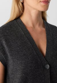 Frau trägt ein dunkelgraues ärmelloses V-Ausschnitt-Strickoberteil mit einem einzelnen schwarzen Knopf, zeigt den Ausschnitt und einen teilweise sichtbaren Anteil des Gesichts mit heller Haut und blonden Haaren.