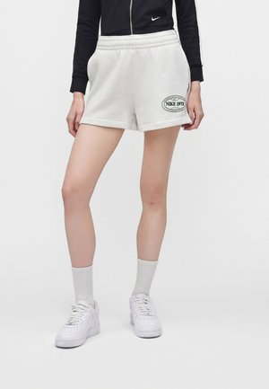 Άτομο που φοράει λευκά σορτς Nike με το λογότυπο "NIKE 1972" στο αριστερό πόδι, λευκές κάλτσες, λευκά αθλητικά παπούτσια και μαύρο μπουφάν με φερμουάρ.