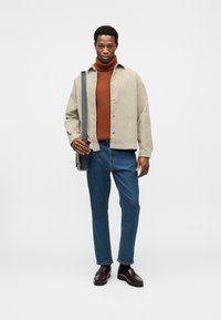 Veste-chemise en velours côtelé beige clair, col roulé rouille, jean bleu, mocassins marron brillants et sac noir ; le mannequin est debout devant un fond uni.