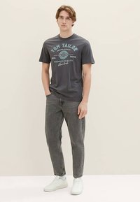 Siva bombažna majica z natisnjenim logotipom, kombinirana s sivimi kavbojkami. Model nosi bele superge. Preprost, priložnostni dizajn.