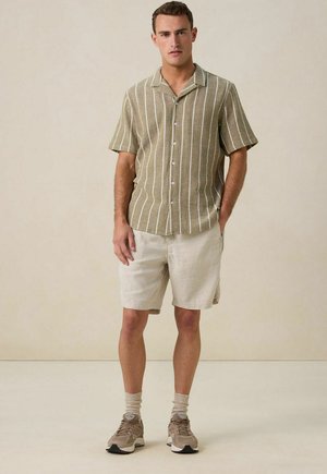 Chemise à manches courtes de couleur vert olive avec des rayures verticales blanches, associée à un short beige clair. Baskets grises et beiges, avec des chaussettes texturées.