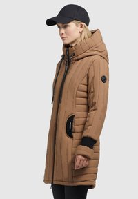 Bruine gewatteerde parka met een hoge kraag, ritssluiting en ribgebreide manchetten. Voorzien van zijzakken met zwarte accenten en een zwarte pet.