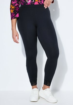 Femme portant des leggings noirs avec des bordures en dentelle et des baskets blanches, debout avec une main reposant sur la hanche.