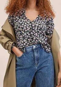 Schwarz mit floralen Mustern, leicht lila und grünen Akzenten, kombiniert mit hoch taillierten blauen Jeans und einem hellbeigen Trenchcoat.