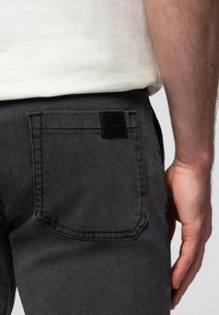 Pantalones de mezclilla gris oscuro con un bolsillo trasero que tiene una etiqueta negra y detalles de costura visibles. Textura suave con un diseño ajustado.