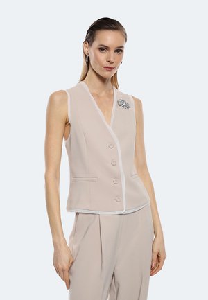 Gilet senza maniche beige chiaro con colletto a sacchetto, bordi bianchi, cinque bottoni e una spilla decorativa sul lato sinistro. Texture liscia.