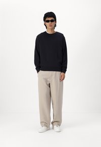 Sort sweatshirt med rund hals, beige brede bukser og hvide sneakers. Model bærer sorte solbriller. Simpelt, minimalistisk design.