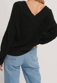 Nainen, joka on pukeutunut mustaan paksuun neuleeseen, jossa on syvä V-muotoinen selkäosa, ja vaaleansinisiin korkeavyötäröisiin farkkuihin, käsi lantiolla.