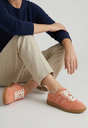 Osoba nosící béžové kalhoty a korálové semišové tenisky s bílými tkaničkami, sedící se zkříženýma nohama na jednobarevném pozadí.