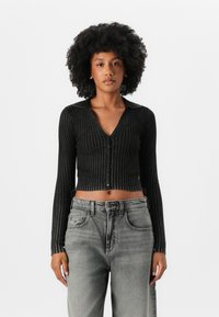 Cárdigan negro con canalé, cuello en V y mangas largas, que cuenta con una fila de botones y un diseño corto. Combinado con jeans grises de talle alto.