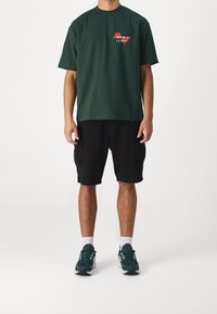 T-shirt in cotone verde con testo stampato rosso, abbinato a pantaloni cargo neri. Sneakers in bianco e nero completano l'outfit.