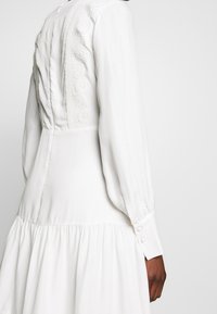 Biała sukienka z gładkiego materiału, z długimi rękawami, dopasowaną górą z koronkowymi detalami oraz rozkloszowanym dołem o marszczonej fakturze.