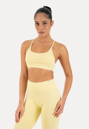Kobieta z ciemnymi włosami związanymi w kok, ubrana w dopasowany bladożółty sportowy biustonosz i legginsy z wysokim stanem, stojąca na białym tle.