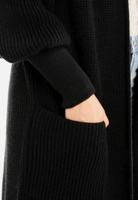 Hand gedeeltelijk in de voorzak van een zwarte gebreide trui gedragen over een witte top en blauwe jeans.