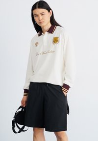 Chemise polo blanche à manches longues avec des rayures bordeaux sur le col et les poignets, présentant un logo et un texte brodés. Short noir avec poches.