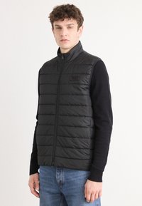 Colete puffer preto com gola alta, textura acolchoada horizontal e fecho frontal. Pequeno logótipo na parte esquerda. Usado por cima de um suéter escuro.