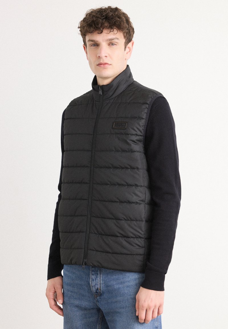 Colete puffer preto com gola alta, textura acolchoada horizontal e fecho frontal. Pequeno logótipo na parte esquerda. Usado por cima de um suéter escuro.