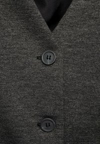 Grijze stof met een gestructureerde uitstraling, met twee zwarte knopen op een kledingstuk. De kraag heeft een V-vorm, wat zorgt voor een gekleed uiterlijk.
