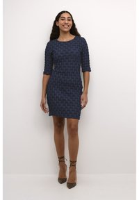 Mörkblå texturerad klänning med vävt mönster, trekvartslånga ärmar och figurnära design, kombinerad med bruna klackar med ankelsnörning.