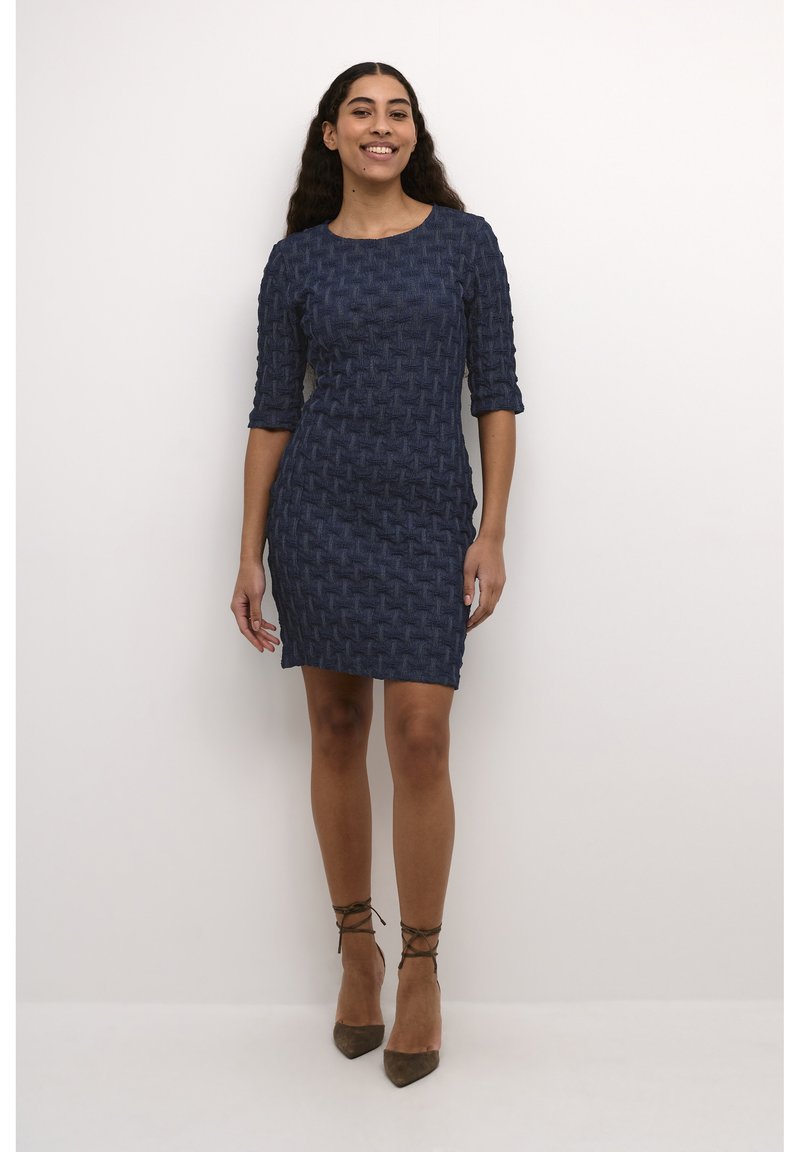 Mörkblå texturerad klänning med vävt mönster, trekvartslånga ärmar och figurnära design, kombinerad med bruna klackar med ankelsnörning.