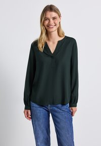 Mörkgrön blus med lång ärm och V-halsringning, mjuk textur, avslappnad passform och rynkade manschetter, i kombination med blå jeans.