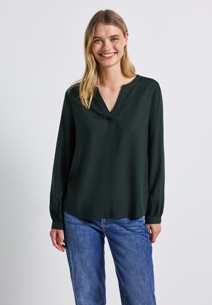 Blusa a maniche lunghe di colore verde scuro con scollatura a V, morbido al tatto, vestibilità rilassata e polsini arricciati, abbinata a jeans blu.