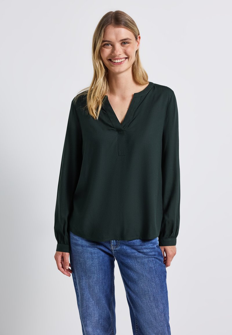 Mörkgrön blus med lång ärm och V-halsringning, mjuk textur, avslappnad passform och rynkade manschetter, i kombination med blå jeans.