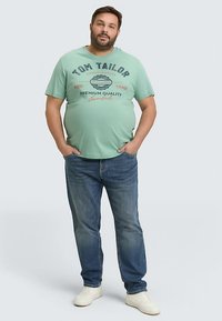 Lichtgroen katoenen T-shirt met bedrukt tekst, gecombineerd met blauwe spijkerbroek en witte sneakers, staand op een effen achtergrond.