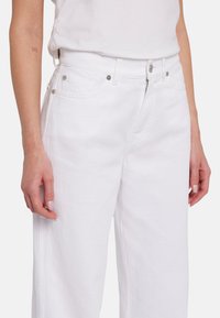 Jeans ampi in denim bianco con vita alta, dotati di tasche anteriori, accessori in metallo argentato e una finitura pulita. Texture liscia, cuciture minime.