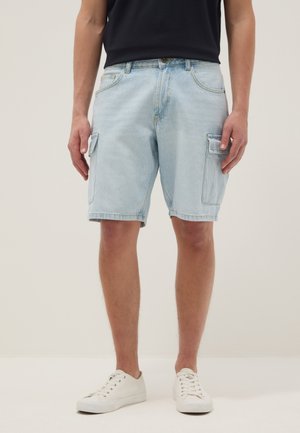 Hombre vistiendo pantalones cortos cargo de mezclilla azul claro con bolsillos con solapa, camiseta negra y zapatillas blancas, de pie frente a un fondo liso.