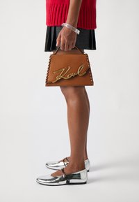 Brown structured handbag with gold script, braided edges; paired with shiny silver flats and a textured black skirt. Clear bracelet on wrist.
