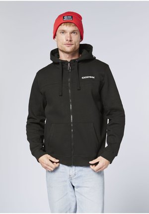 Black zip-up hoodie with a drawstring hood, kangaroo pockets, and a white logo on the chest. Worn with light-colored jeans and a red beanie.