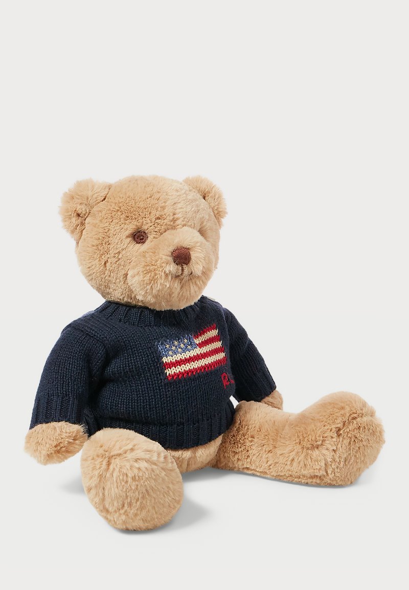 Urso de peluche castanho vestindo um suéter de malha azul-marinho com um design da bandeira americana, sentado com patas arredondadas e textura de pelúcia suave.