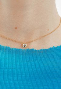 Collar de cadena de oro con un pequeño colgante de gema facetada y redonda. Lleva puesto sobre una blusa texturizada de un azul brillante con un escote deshilachado.