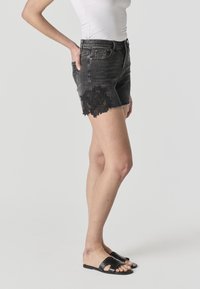 Donna con pantaloncini di jeans neri con dettagli in pizzo floreale sul lato, canotta bianca e sandali neri a ciabatta, in piedi di profilo.