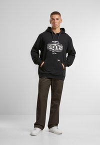 Black hoodie with front pocket and white "DICKIES" logo, paired with dark brown pants and white sneakers on a plain background.