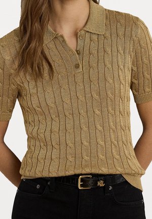Femme portant un polo à manches courtes beige en maille torsadée, rentré dans un pantalon noir avec une ceinture noire avec boucle dorée.